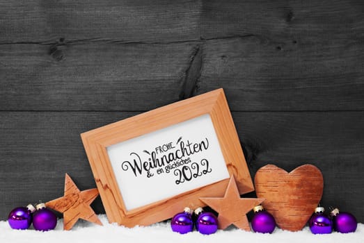 Frame With German Calligraphy Frohe Weihnachten Und Ein Glueckliches 2022 Mean Merry Christmas And A Happy 2022. Purple Christmas Decoration Like Ball, Star And Heart. Gray Wooden Background With Snow