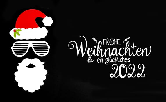 German Und Ein Glueckliches 2022 Means Merry Christmas And A Happy 2022. Santa Claus Paper Mask With Christmas Decoration And Accessories. Black Paper Background