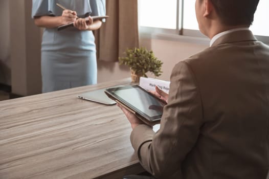 Businessman manager in brown suit using PC tablet while sitting at office desk and secretary in blue dress taking notes while standing in front of his boss. Business work concept