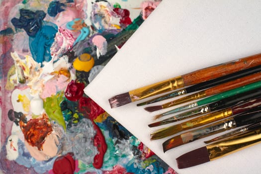 top view closeup of artist mixing palette with colored painting brushes and white empty canvas