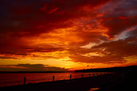 Red cloudy sunset above lake at dusk. Bloody sky above bridge and reflective water surface waves. Evening cloudscape in twilight. Ruddy horizon sky and river. Morning fiery orange sunrise in summer