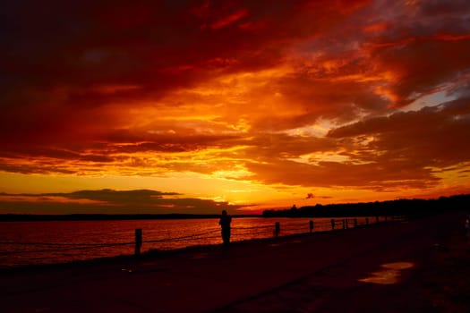 Lonely man silhouette watches red cloudy sunset above lake at dusk. Sad boy on the bridge stands under sky and reflective water surface. Evening cloudscape in twilight. Fiery orange sunrise in summer