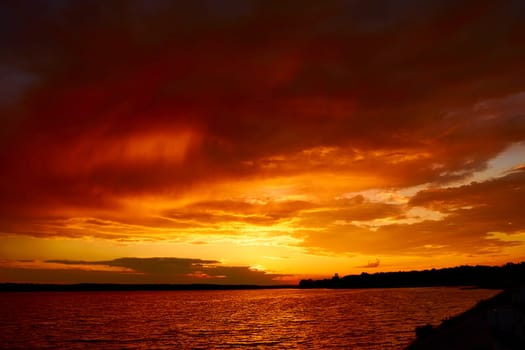 Red cloudy sunset above lake at dusk. Bloody sky above reflective water surface waves in wind. Evening cloudscape in twilight. Ruddy horizon sky and river. Morning fiery orange sunrise in summer dawn