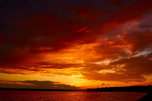 Red cloudy sunset above lake at dusk. Bloody sky above reflective water surface waves in wind. Evening cloudscape in twilight. Ruddy horizon sky and river. Morning fiery orange sunrise in summer dawn