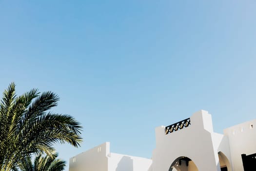 Bottom view of palm trees, resort hotel white buildings and blue clear sky. Hotel business concept. Summer or winter vacation trip. Family relaxation time. High quality photo