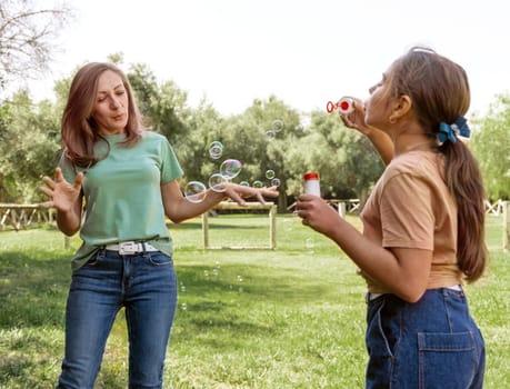 Mother and daughter blowing soap bubbles outdoors. Cute kid girl and middle aged woman having fun in a park in summer. Children outdoor activity concept, authentic childhood moment