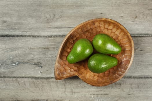 Table top view, three whole avocados in wooden carved bowl placed on gray wood desk.