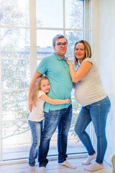 Beautiful pregnant woman, girl and man standing near the window