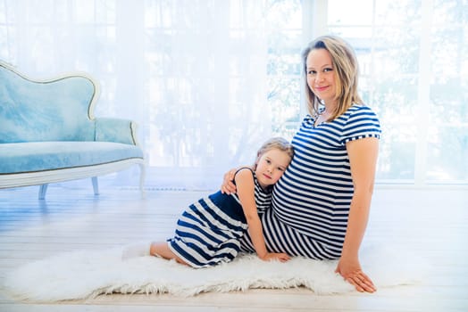 Beautiful pregnant woman with daughter sitting near window at home