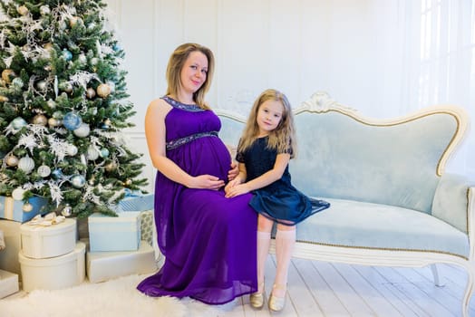 Beautiful pregnant woman in an ultraviolet dress sitting with daughter on the sofa near christmas tree