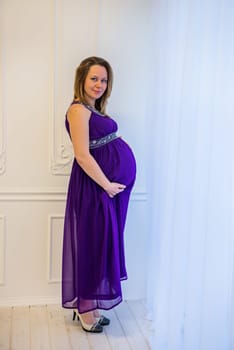 Beautiful pregnant woman in an ultraviolet dress standing near the window.