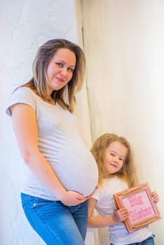 Beautiful pregnant woman with daughter standing near wall at home.