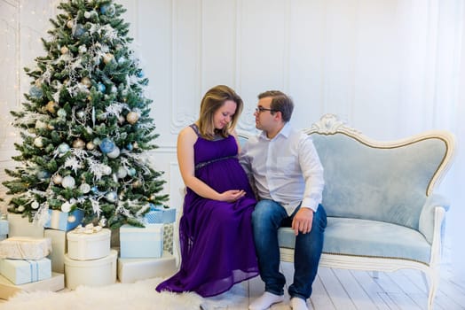 Pregnant woman in an ultraviolet dress with husband sitting on the sofa near Christmas tree