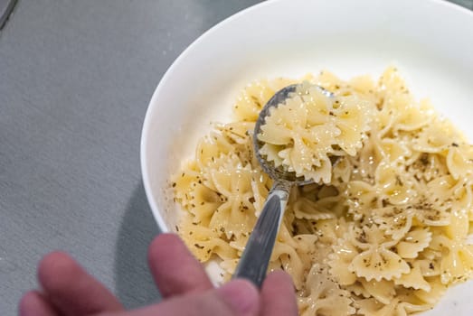 Step by step home cooking salad recepie. The view is close to the hand holding a spoon against the white bowl with farfalle paste