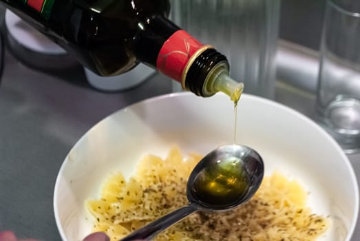 Step by step home cooking salad recepie. Olive oil pours into a spoon against the background of a bowl with pasta
