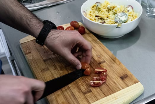 Step by step home cooking salad recepie. A man cuts cherry tomatoes on a wooden board against a white bowl of pasta