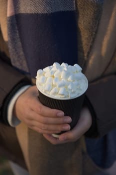 Vertical image of young and youthful female hands holding a hot chocolate to go in a black and white paper cup. Earthy color winter clothes background
