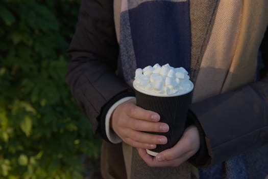 Warm evening sun highlights marshmallows atop of hot takeaway cacao drink. Held by a young women wearing winter clothes outdoors, dark leaf background