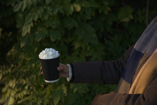 Young women in winter coat and scarf holds a takeaway beverage, hot chocolate, topped creamy marsh mallows. Golden outdoor light, leafy background