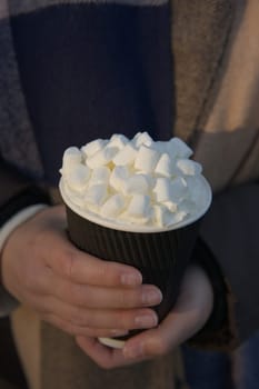 Hot take away beverage with an abundance of marshmallows and cream on top, cradled by youthful female hands. Likely hot chocolate, coffee or tea.