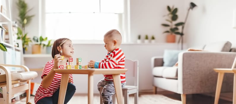 Sister teaching little brother letters by using cubes with symbols indoors. Concept of the family relationship.