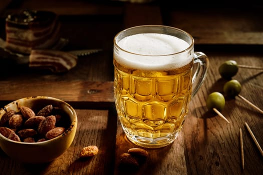 Glass of cold refreshing beer placed on wooden table with salted almond in bowl and green olives on toothpicks