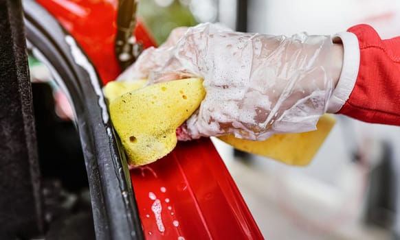 Young woman cleaning rear part of her car, closeup detail on hand in glove holding yellow sponge.