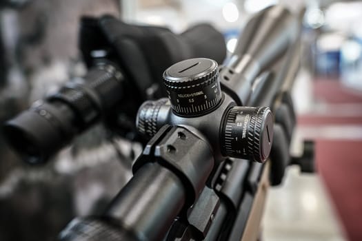 Black shooting scope optics mounted on rifle displayed at weapons exhibition fair, closeup detail to adjustment knobs.