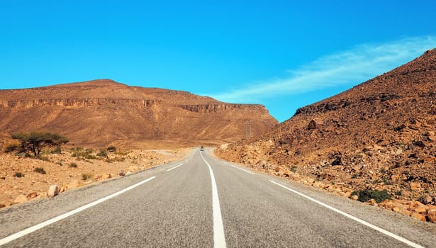 Straight asphalt road , small Atlas mountains and low bushes both sides, clear sky above - typical landscape in Southern Morocco.