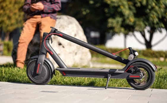 Folded electric scooter on concrete pavement, young man legs in background.