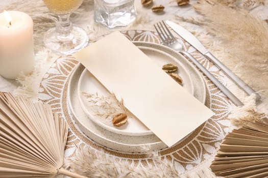 Table place with blank card on white wooden table with bohemian decorations around close up. Boho wedding menu card mockup. Tropical beige template with dried palm leaves and pampas grass, place fot text