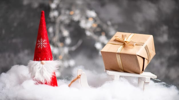 Christmas gnome greeting card with bokeh lights and snow, winter season, gift on the sledge