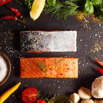 Fresh uncooked salmon fillets placed one up, one down, in a frame of ingredients for cooking the meal including tomatoes, garlic, chili peppers, herbs and seasoning, square format overhead view