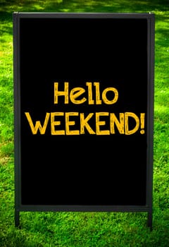 HELLO WEEKEND  message on sidewalk blackboard sign against green grass background. Copy Space available. Concept image