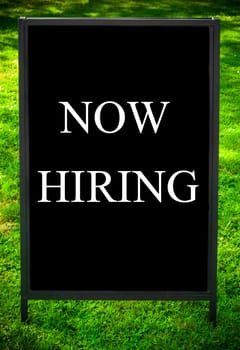 NOW HIRING  message on sidewalk blackboard sign against green grass background. Copy Space available. Concept image