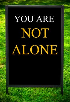 YOU ARE NOT ALONE  message on sidewalk blackboard sign against green grass background. Copy Space available. Concept image
