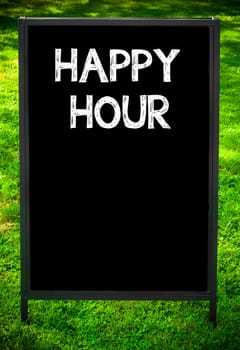 HAPPY HOUR  message on sidewalk blackboard sign against green grass background. Copy Space available. Concept image