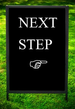 NEXT STEP message and hand pointing to the right on sidewalk blackboard sign against green grass background. Copy Space available. Concept image
