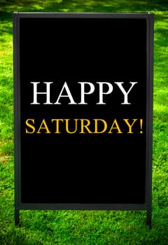 HAPPY SATURDAY  message on sidewalk blackboard sign against green grass background. Copy Space available. Concept image