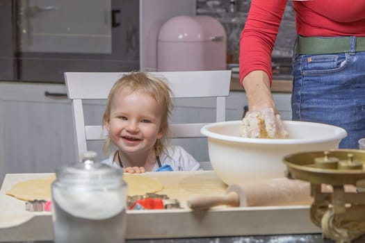 Little chef. Cute little boy is laughing at the camera while his mom knead the dough in domestic kitchen.