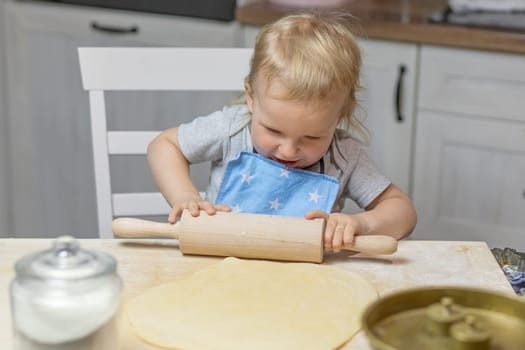 Little helper boy is rolling dough with a wooden rolling pin in domestic kitchen, indoors. Horizontally.