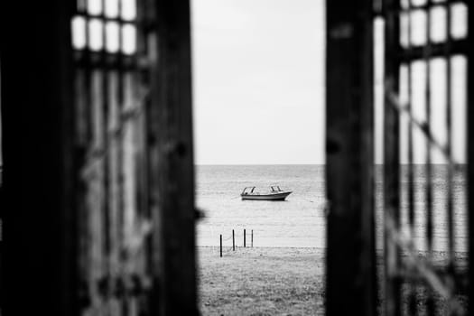 Black-white vessel or yacht photography. Opened doors to beach with alone boat or ship in silent sea or ocean water surface. Window opening to quiet and peaceful harbor or bay. No waves tranquil mood