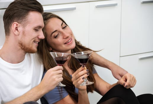 Smiling man and woman sitting on kitchen floor and holding glasses of cocktails. Romantic couple spending time together, having fun and enjoy drinking at home.