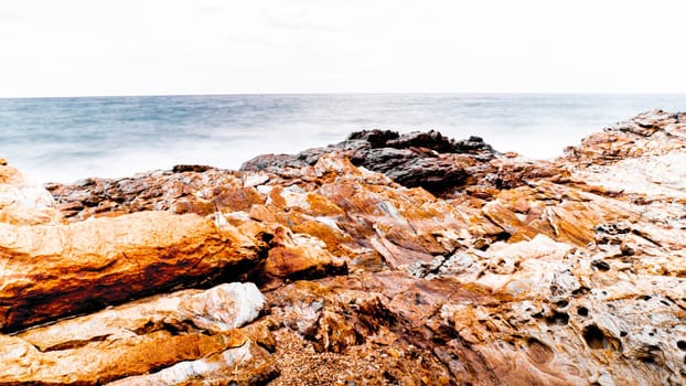 Scenic Layered Rocks in blue sea against sky with forest hills background. Sea Waves Splash on Rock