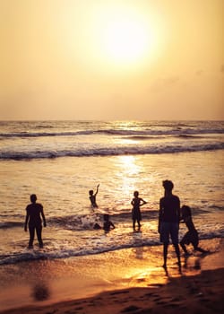 Kalutara, Sri Lanka - April 15, 2017: Silhouettes of people, mostly kids playing on the wet beach, sea and golden sunset afternoon light in back.