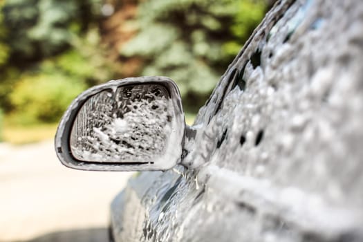 Side mirror of silver car, covered with shampoo and foam, when washed in carwash on a sunny day.