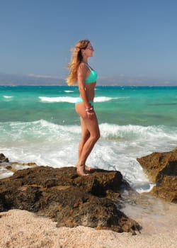 Young athletic woman with bubble butt, wearing cyan blue bikini ad sunglasses, standing on rock beach with turquoise sea in background.