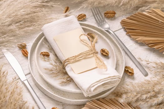 Table place with blank card on white wooden table with bohemian decorations around close up. Boho wedding menu card mockup. Tropical beige template with dried palm leaves and pampas grass, place fot text