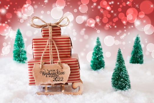 Sled With Christmas Gift And Label With German Frohe Weihnachten Und Ein Glueckliches 2022 Means Merry Christmas And A Happy 2022. Snow With Christmas Tree And Red Sparkling Background