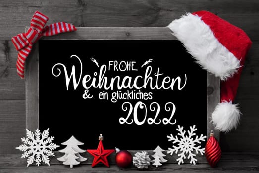 Chalkboard With German Calligraphy Frohe Weihnachten Und Ein Glueckliches 2022 Means Merry Christmas And A Happy 2022. Christmas Decoration Like Tree, Ball And Santa Hat. Gray Wooden Background